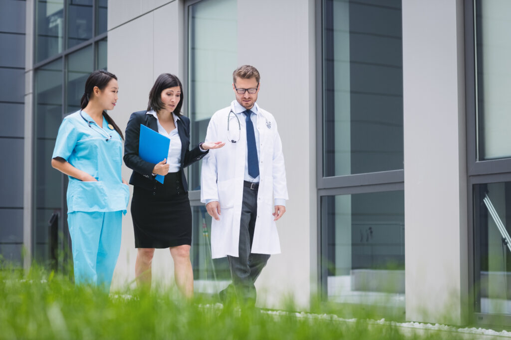 Healthcare professionals and a businesswoman walking outside a modern medical building, discussing documents and patient care.