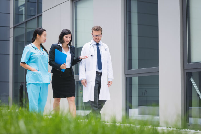 Healthcare professionals and a businesswoman walking outside a modern medical building, discussing documents and patient care.