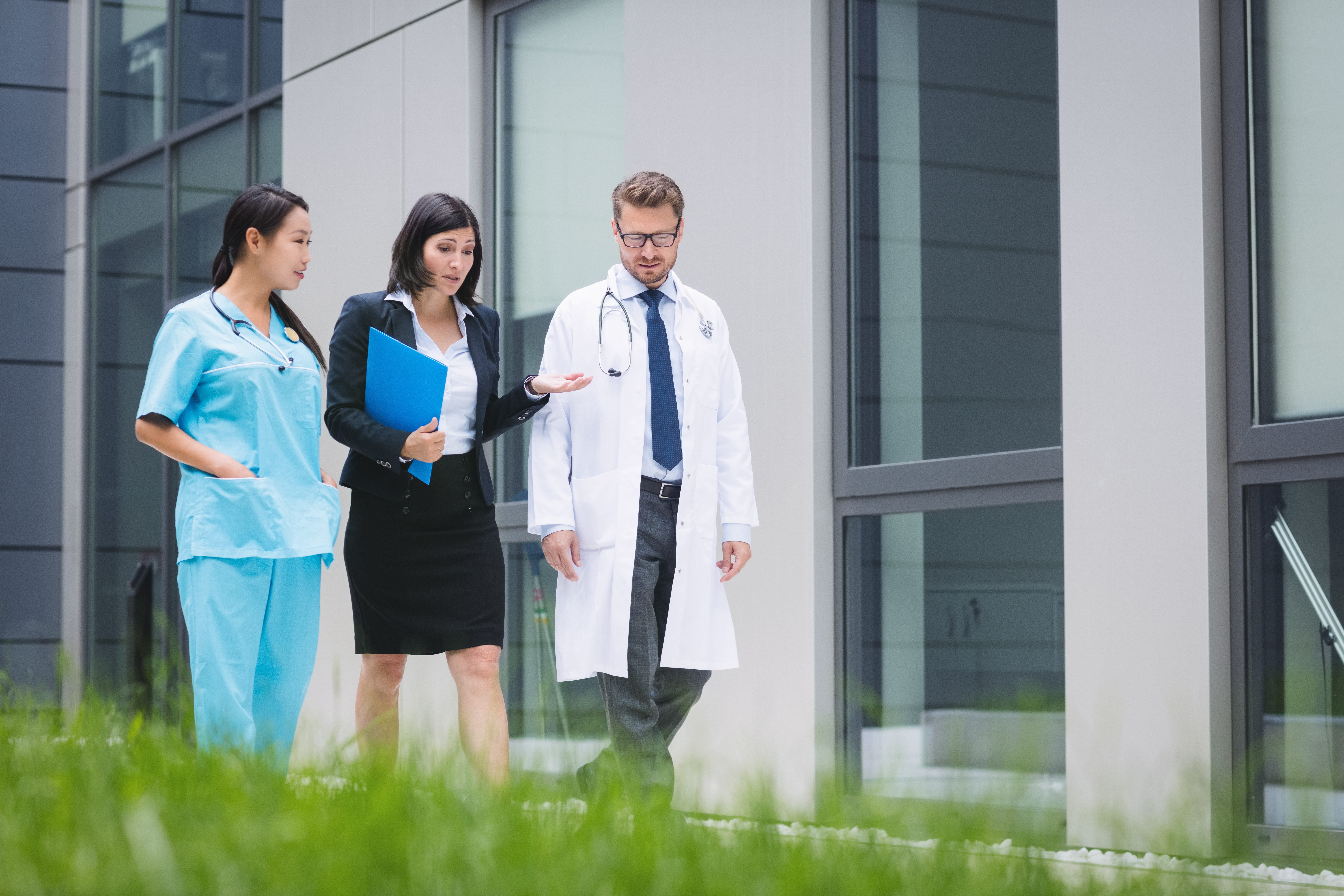 Healthcare professionals and a businesswoman walking outside a modern medical building, discussing documents and patient care.