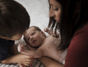 A mother holding a newborn baby while their brother watches.