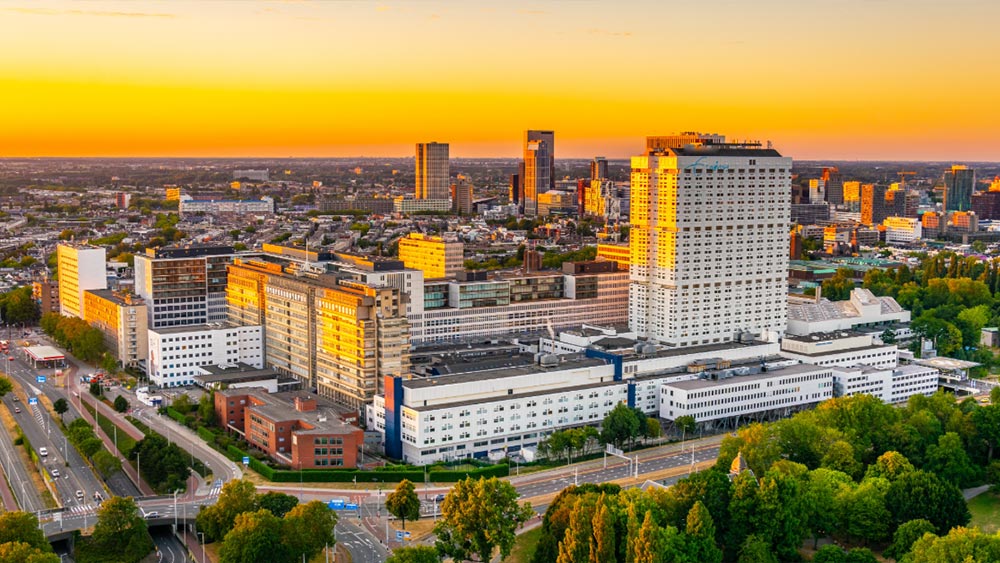 Aerial view of a large hospital complex in an urban setting at sunset, surrounded by city buildings, roads, and green space.